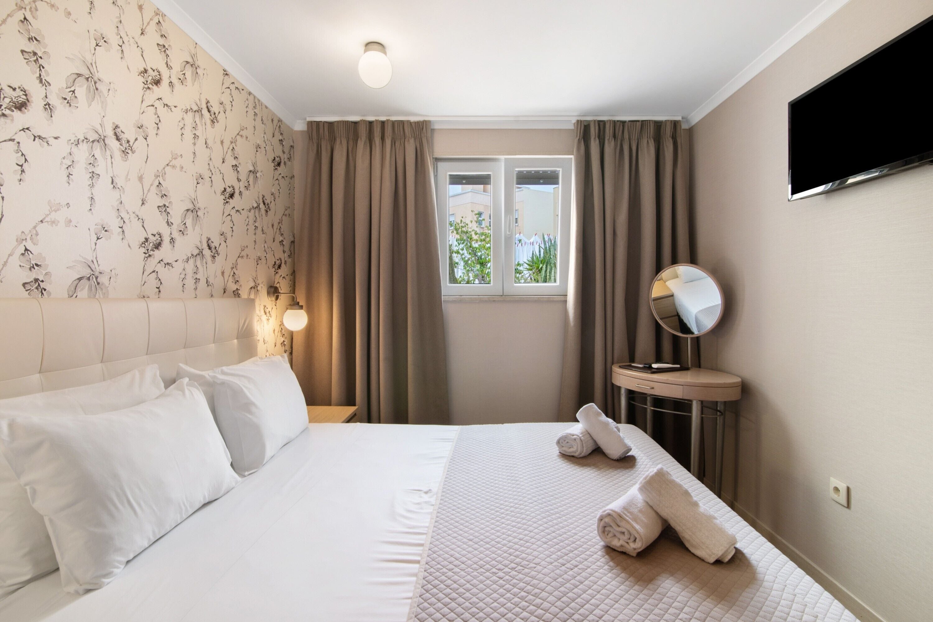 Une chambre d’hôtel avec un lit, des rideaux, une télévision, un miroir rond et une fenêtre donnant sur un paysage.