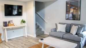 Una sala de estar con un sofá gris, una mesa de centro blanca, un televisor montado en la pared y una escalera al fondo.