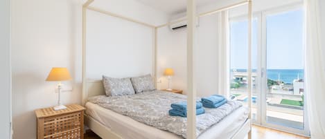 A four-poster bed with a patterned bedspread, two bedside lamps, a wicker side table, and a view of the sea through large sliding glass doors.