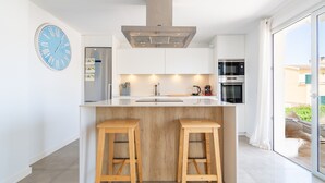 Une cuisine moderne dotée d’un îlot central, de tabourets en bois, d’un réfrigérateur en acier inoxydable et d’une grande fenêtre.