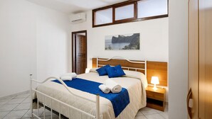 A neatly made bed with a blue blanket and pillows, a bedside table with a lamp, a picture of a coastal landscape, and a window with a view of the sea.