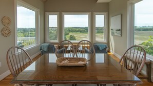 A dining room with a large wooden table, four chairs, and a view of a green landscape through large windows. - Tequila Sunset:Amazing Dog Friendly Home;Ocean Views/Private Dock; Minutes to Beach;Elevator/WiFi (Edisto Island)