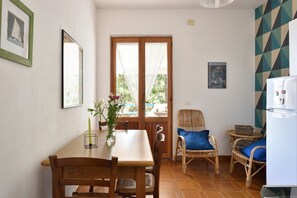 Un comedor con una mesa de madera, sillas, un florero con flores, una ventana con cortinas, una silla de mimbre con un cojín azul y un cartel en la pared.