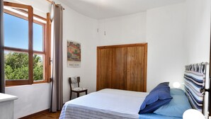 Ein Schlafzimmer mit einem Bett, einem Kleiderschrank, einem Sessel, einem Schreibtisch, einer Lampe und einem Fenster mit Vorhängen.