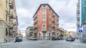 A multi-story building with a corner entrance, parked cars, and a pedestrian crossing. - GuestHost - Torino Old Fashion Apartment with Balcony (Torino)