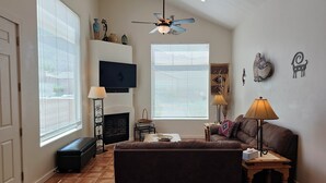 A living room with a fireplace, a television mounted above it, a brown sofa, a small wooden table with a lamp, and a large window with blinds. - Moab Rim Village 2BR condo w/ pool & red rock view (Moab)