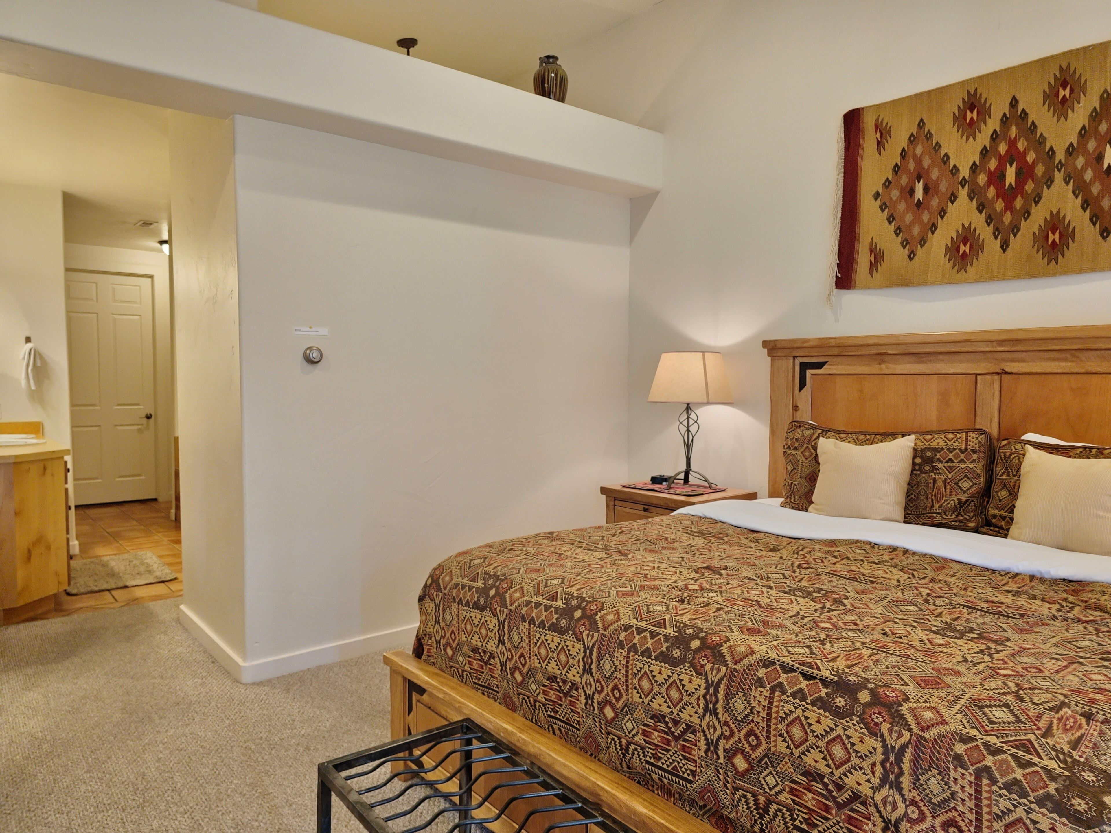 A bedroom with a large bed, a wooden headboard, a nightstand with a lamp, and a patterned wall hanging.