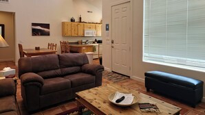 A cozy living room with a brown leather couch, a wooden coffee table with a remote and papers, a dining area with a wooden table and chairs, a kitchen with wooden cabinets and a microwave, and a large window with blinds. - Moab Rim Village 2BR condo w/ pool & red rock view (Moab)