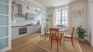 Une cuisine moderne avec des armoires blanches, un four encastré et des luminaires suspendus. On y trouve un coin repas avec une table ronde et des chaises, ainsi qu’une plante en pot.