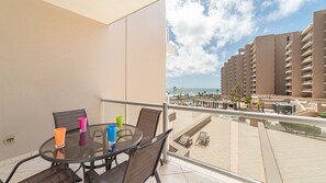 Un balcón con una mesa y sillas de vidrio, con vistas a una zona de piscina y un hotel frente a la playa.