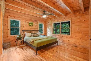 A wooden cabin bedroom with a bed, two windows, a ceiling fan, and a nightstand.