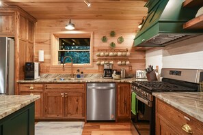 A kitchen with wooden cabinets, a stainless steel dishwasher, a built-in oven, a gas stove, and a window with a view of greenery. - The Leaflet | Hocking Hills, Swim Spa, Theater (Laurelville)