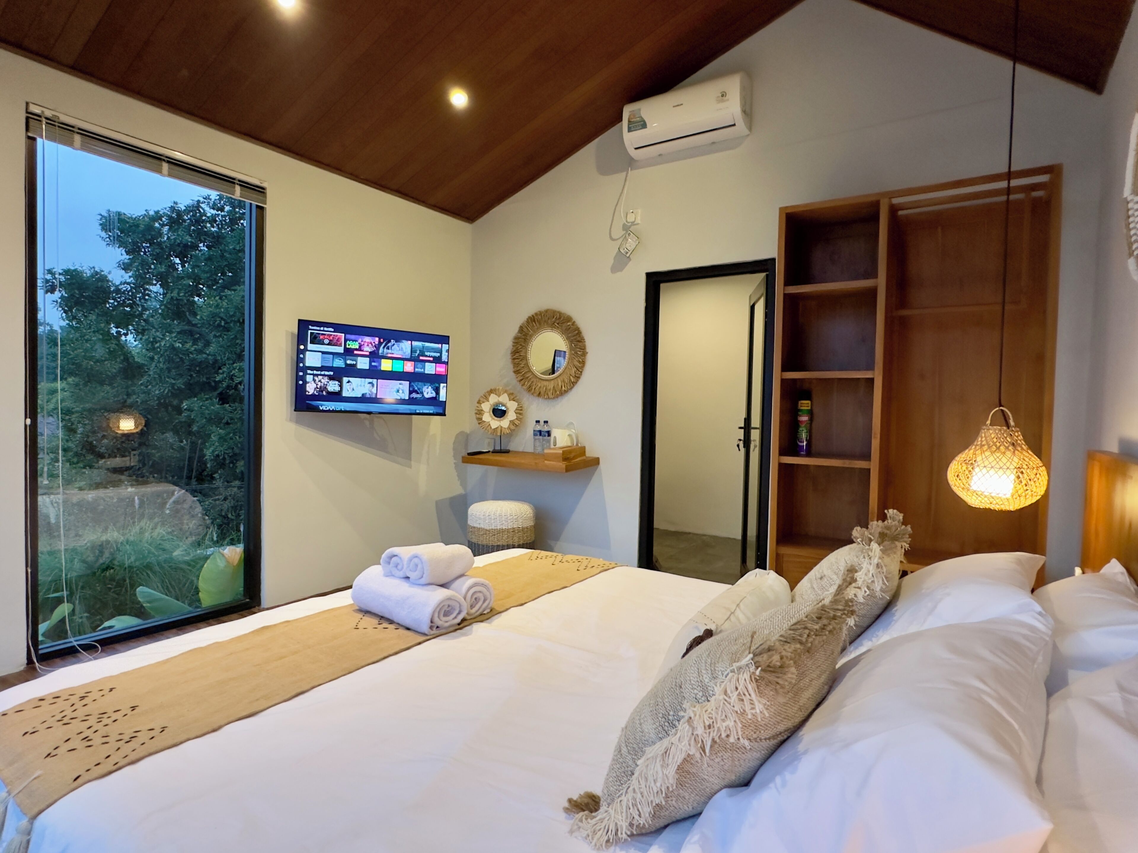 A modern bedroom with a large bed, a TV on the wall, a wooden shelf, and a view of greenery outside.
