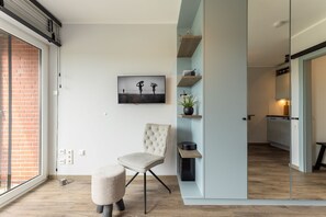Een moderne woonkamer met een groot raam, een comfortabele stoel, een klein voetenbankje en een minimalistische inrichting, waaronder een ingelijst kunstwerk en planken met decoratieve objecten.