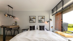 Ein modernes Schlafzimmer mit einem großen Bett, einem Schreibtisch mit zwei Stühlen und Blick auf einen Garten durch ein großes Fenster.
