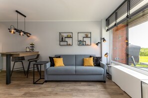 Een moderne woonkamer met een grijze bank, twee gele kussens, een houten eettafel met twee stoelen, een hanglamp en een raam met uitzicht op een bakstenen muur en begroeiing.