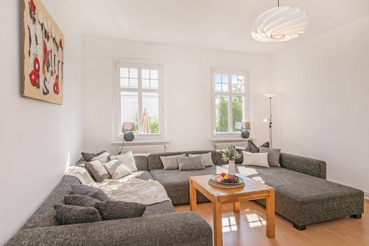 Een moderne woonkamer met een hoekbank, een houten salontafel en twee ramen met witte kozijnen.