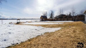 A residential area with a wooden cabin, a fenced yard, and a body of water in the background. - Modern Lake Lucille Lakefront, Whole 5 bedroom house, Private dock & games
 (Wasilla)