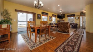 A spacious living room with a wooden dining table, chairs, a sofa, and a TV. There is a large window providing a view of the outdoors. - Modern Lake Lucille Lakefront, Whole 5 bedroom house, Private dock & games
 (Wasilla)