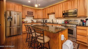 A kitchen with wooden cabinets, stainless steel appliances, a granite countertop, and bar stools. - Modern Lake Lucille Lakefront, Whole 5 bedroom house, Private dock & games
 (Wasilla)