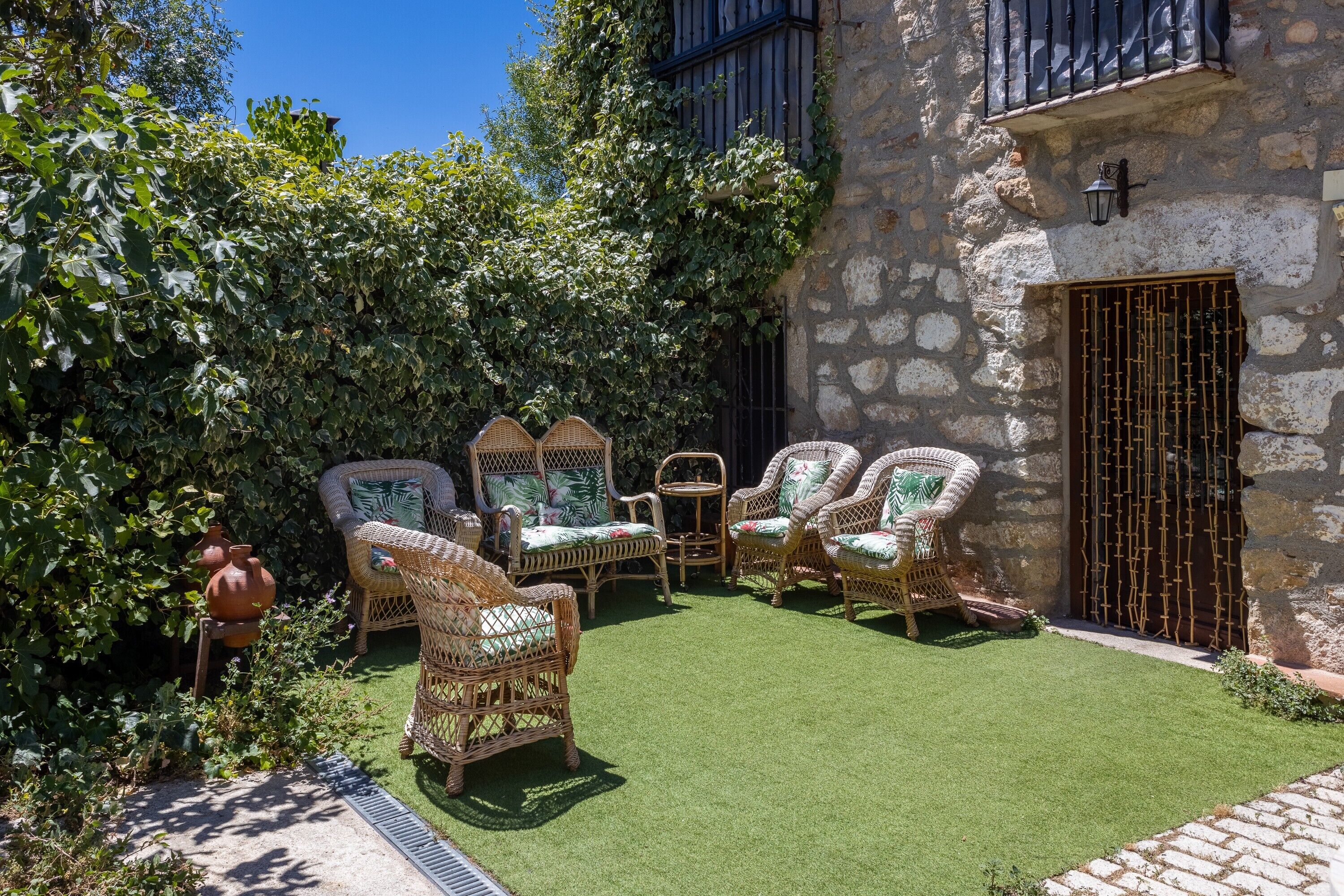 Un edificio de piedra con una reja de hierro forjado, un pequeño patio con césped artificial, muebles de mimbre y una planta en maceta.
