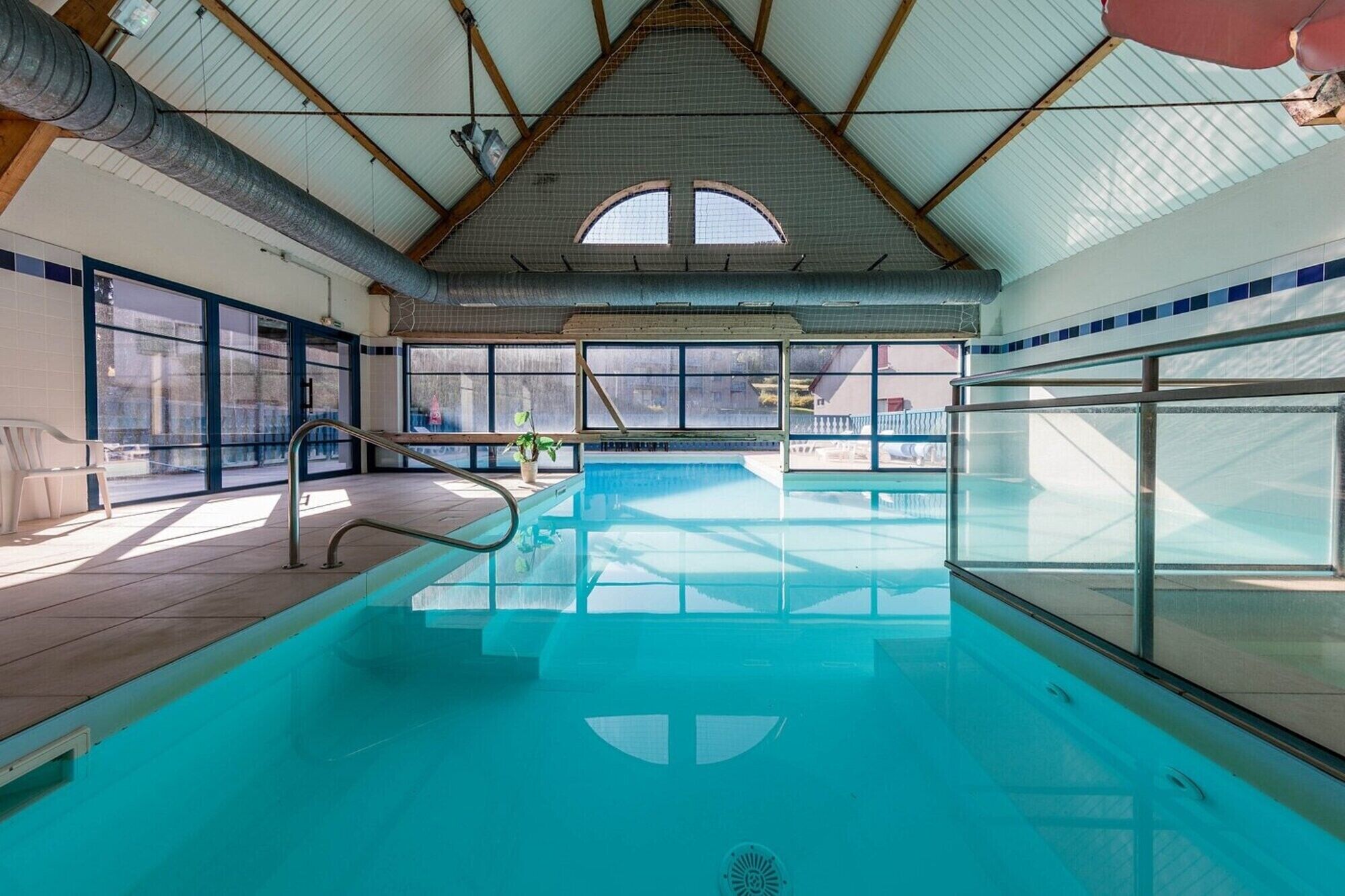 Una piscina interior amb parets de vidre transparent, un paviment de rajoles i un sostre alt amb bigues estructurals visibles.