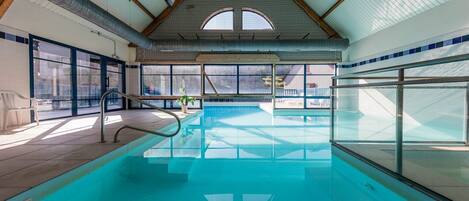 An indoor swimming pool with clear glass walls, a tiled floor, and a high ceiling with visible structural beams.