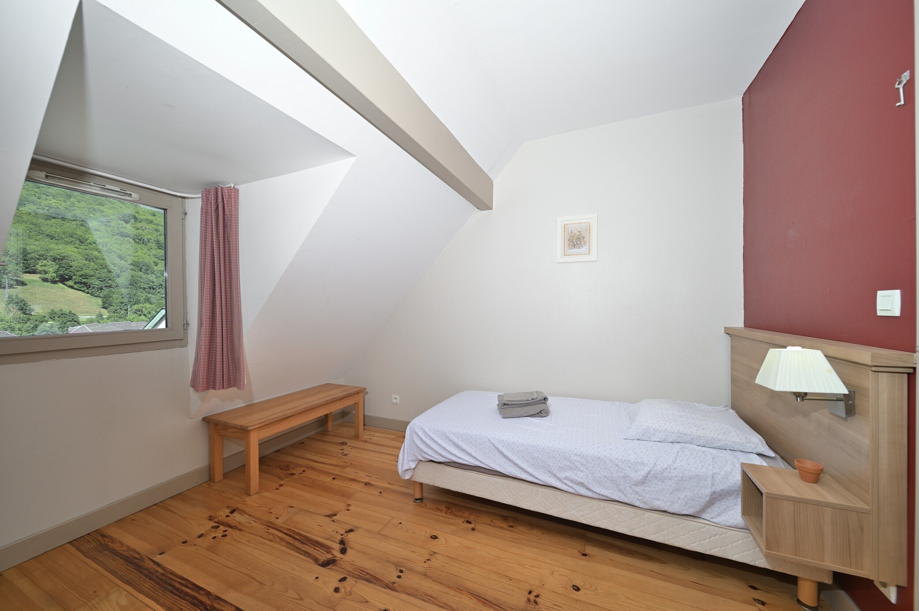 Une chambre avec un plafond en pente, un lit, un banc, une fenêtre donnant sur un beau paysage et une petite table de chevet.