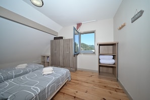 Une chambre avec deux lits, une armoire, une étagère avec des serviettes et une fenêtre donnant sur des collines.