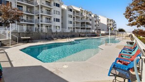 A swimming pool area with a clear blue pool, lounge chairs, and a multi-story building with balconies.