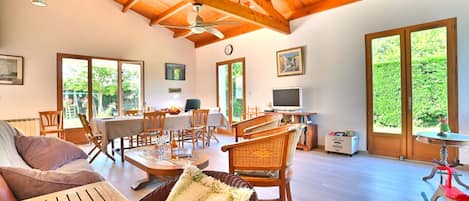 A spacious living area with a high ceiling, wooden beams, a dining table, wicker furniture, and large windows offering a view of greenery.