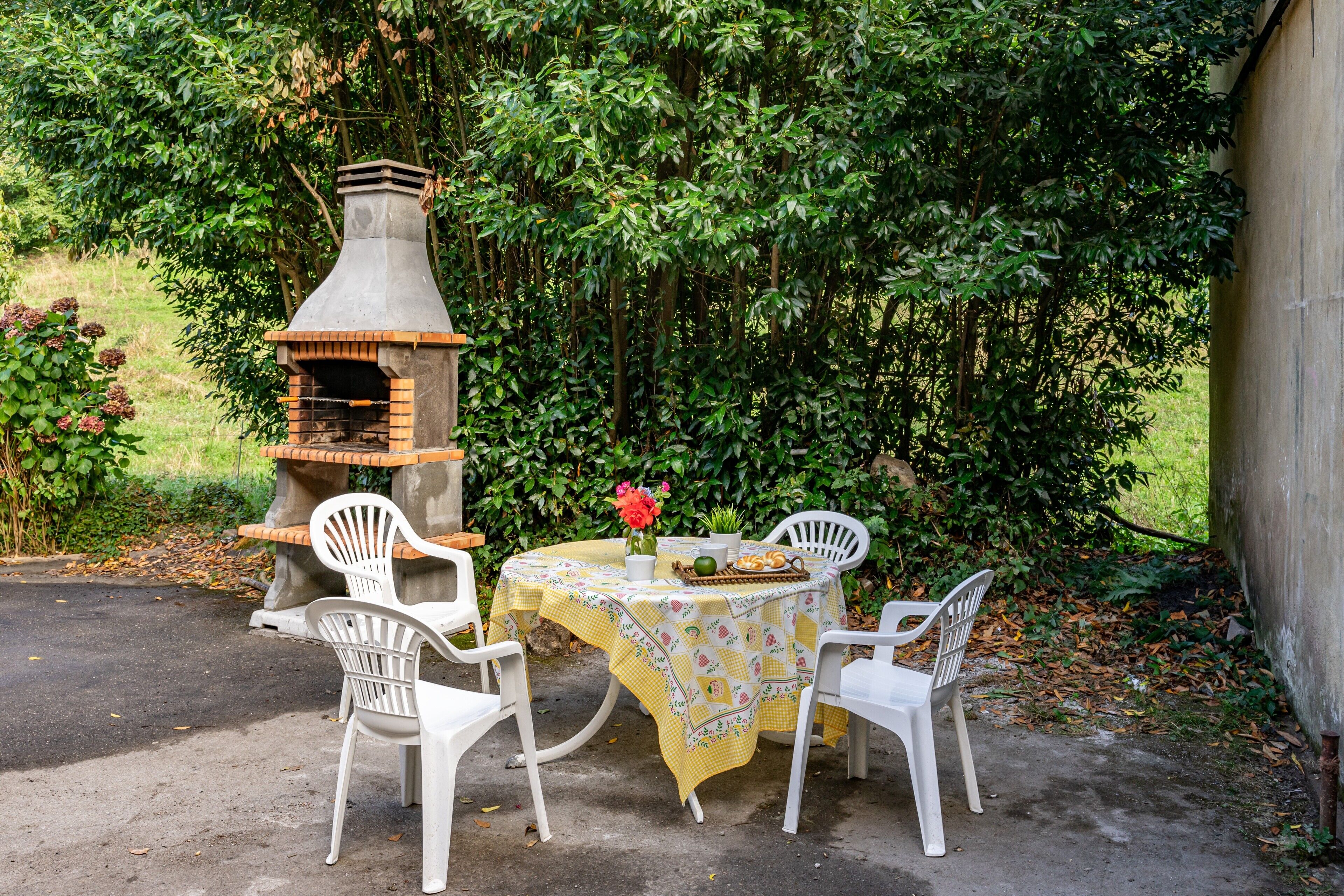 Une terrasse extérieure avec un four en briques, des chaises en plastique blanches et une table recouverte d’une nappe jaune.