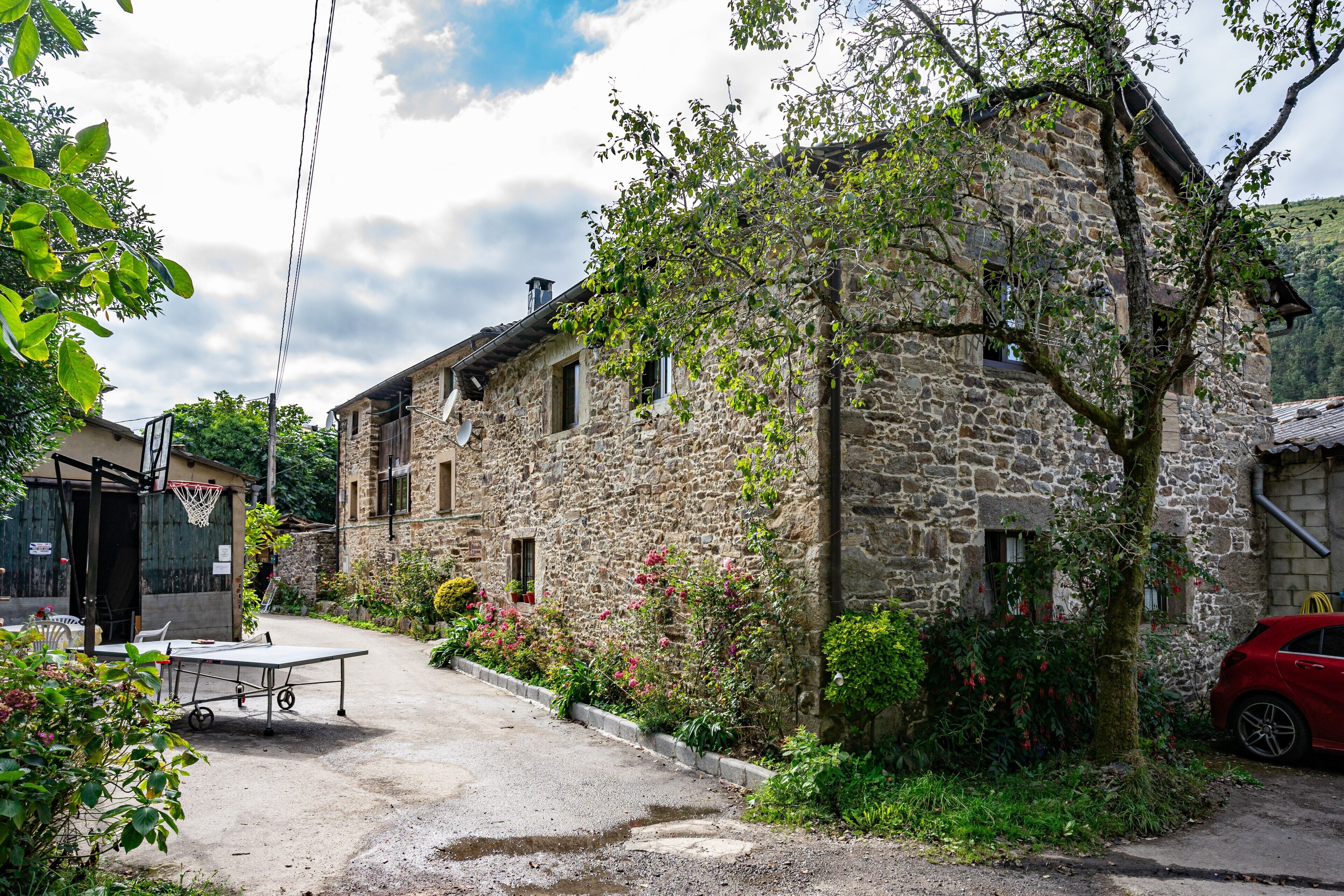 A stone building with a basketball hoop, a red car, and a tennis table in the foreground.