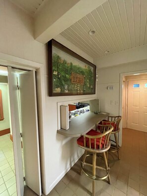 Uma área de refeições com uma mesa branca e quatro cadeiras de madeira, um quadro sobre a mesa e uma cozinha com fogão e pia.