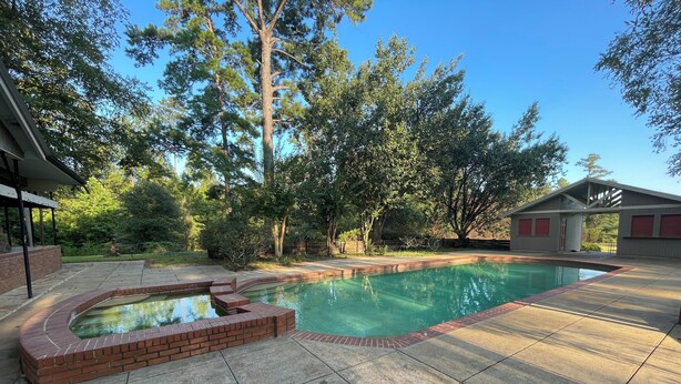 A backyard with a swimming pool, a brick hot tub, and a covered patio area. - One-of-a-Kind Railcar Retreat with Pool (Tylertown)