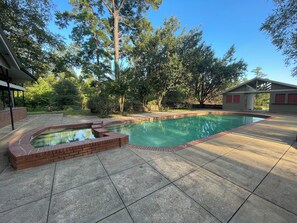A backyard with a swimming pool, a brick hot tub, and a covered patio area. - One-of-a-Kind Railcar Retreat with Pool (Tylertown)