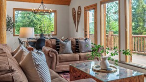 A cozy living room with a brown sofa, a glass coffee table, and a view of a wooden deck through large windows.