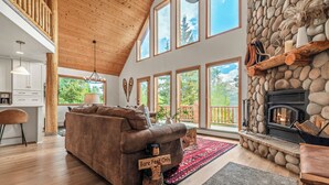 A spacious living room with a stone fireplace, a sofa, and large windows offering a view of the outdoors.