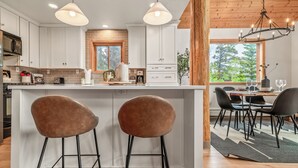 A modern kitchen with white cabinets, a central island with stools, and a dining area with a view of trees.