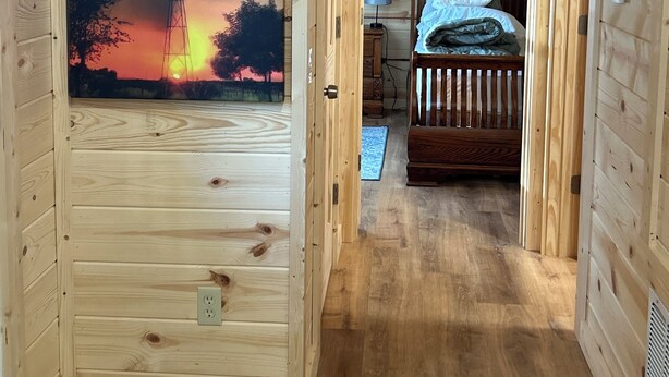 A wooden cabin interior with a large window displaying a sunset view, a bed with a headboard, and a bedside table.