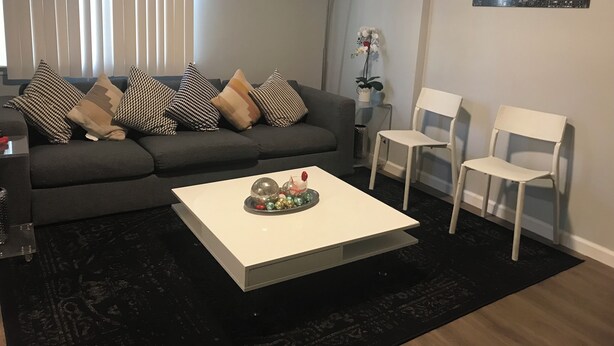 Ett modernt vardagsrum med en grå soffa, vita stolar, ett vitt soffbord och en tavla med en stadsbild på väggen.