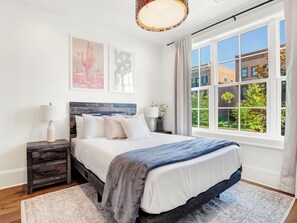 A bedroom with a large window, a bed with white bedding, a nightstand, and a wall-mounted lamp.