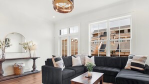 A modern living room with a black sectional sofa, a wooden coffee table, and a large window offering a view of a brick building. - Luxe New Construction with River View, Pool Table (Savannah)