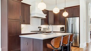 A modern kitchen with dark wood cabinets, a stainless steel refrigerator, a central island with bar stools, and pendant lighting. - Luxe New Construction with River View, Pool Table (Savannah)