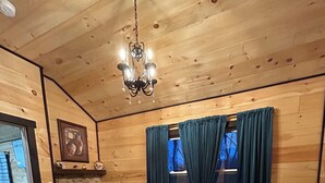 A cozy wooden room with a bed, a fireplace, a chandelier, and a window with curtains. - Gorgeous New Cabin with 70Acres!Trails, Waterfalls and Amazing Rock Formations! (Cub Run)