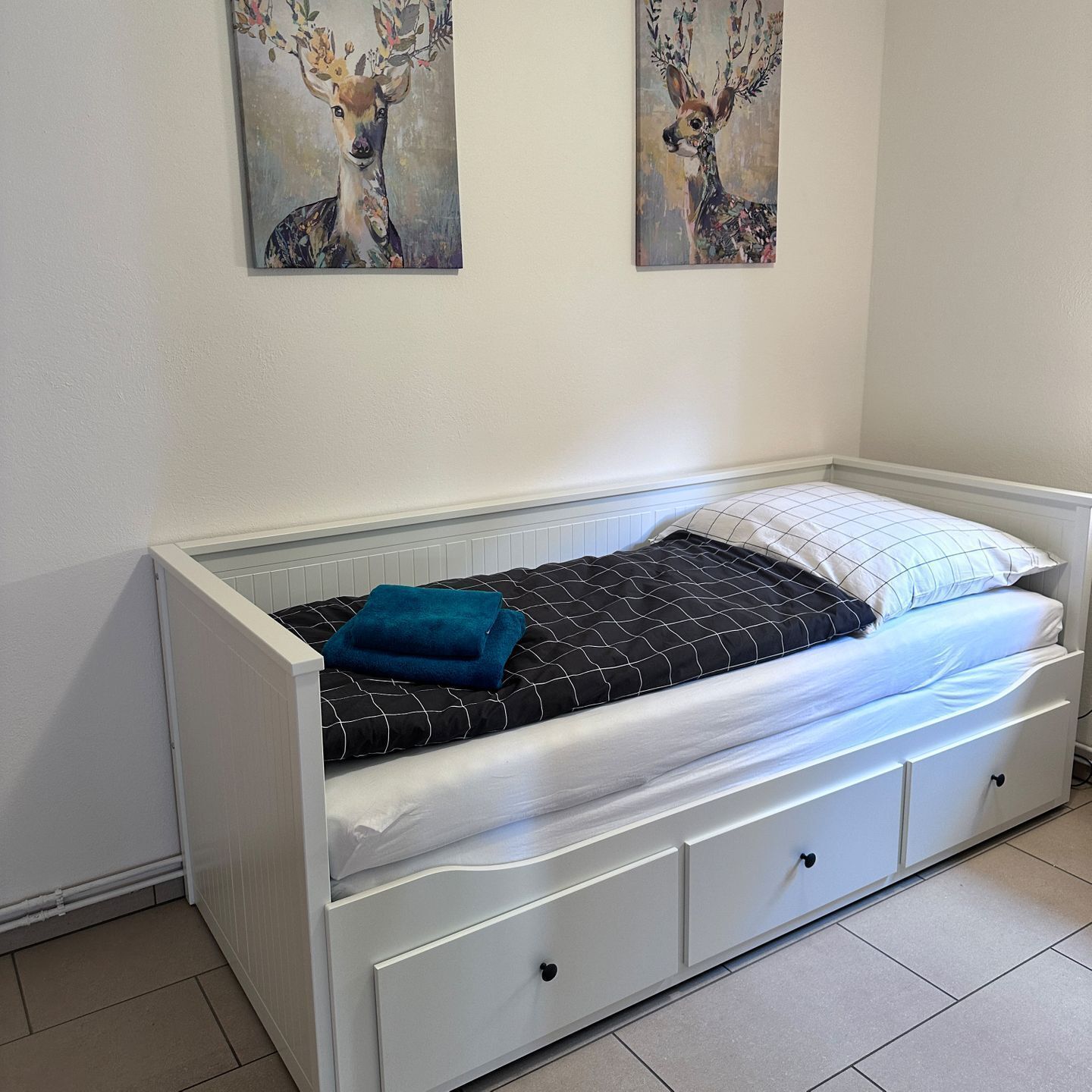 A single-bed room with a white headboard, a black and white patterned bedspread, a blue pillow, and a white pillow. There are two wall-mounted paintings of deer above the bed.