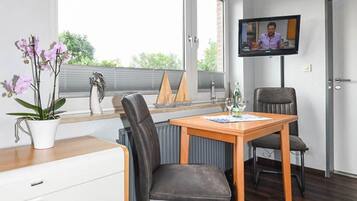 Ein modernes Zimmer mit einem Holztisch, zwei Stühlen, einem Fernseher, einer Topfpflanze und einem Fenster mit Jalousien.