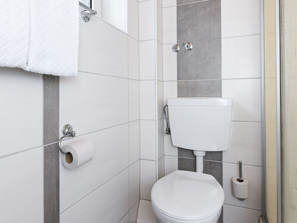 A small, modern bathroom with white tiles, a toilet, a towel rack, and a toilet paper holder.