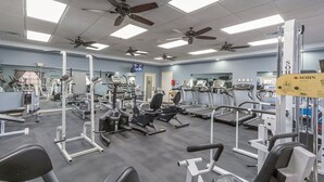 Een sportschool met diverse fitnessapparaten, plafondventilatoren en een televisie aan de muur.