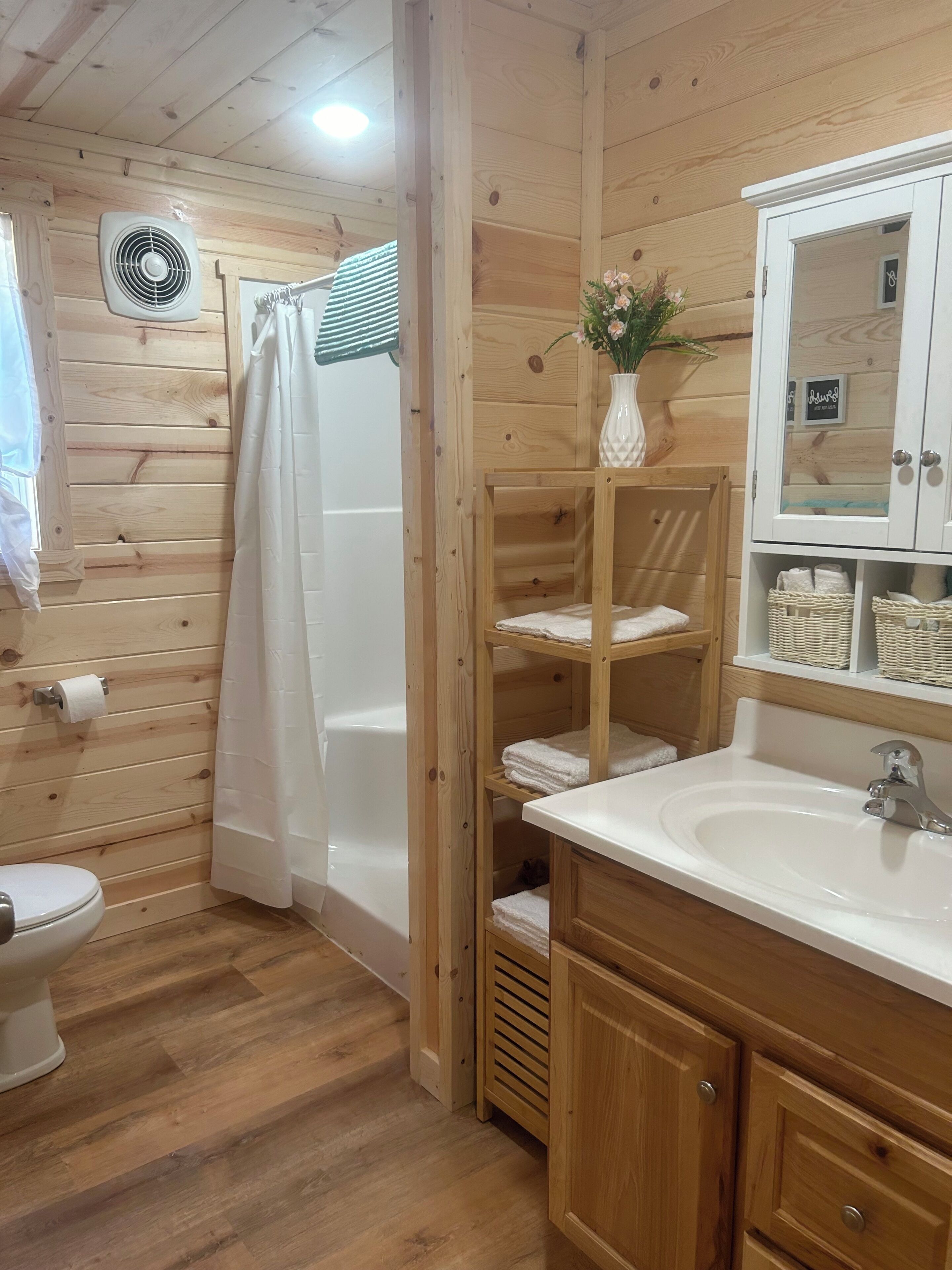 A wooden-paneled bathroom with a toilet, shower, sink, and shelves with towels.